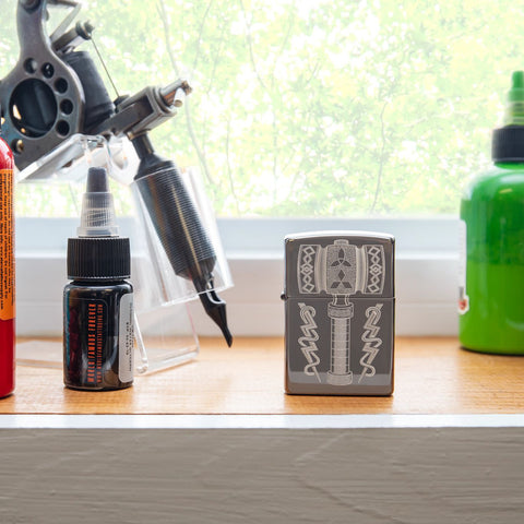 Lifestyle image of Thor's Hammer Design Black Ice® Windproof Lighter standing on a window sill with a tattoo gun and ink bottles